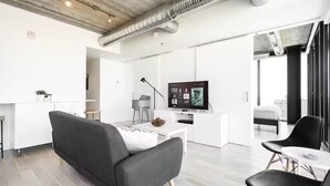 Een moderne woonkamer met een grijze bank, een kleine witte tafel en een televisie. Er is een keuken met een wit aanrecht en een barkruk. De kamer heeft een hoge plafond met zichtbare ventilatiekanalen.