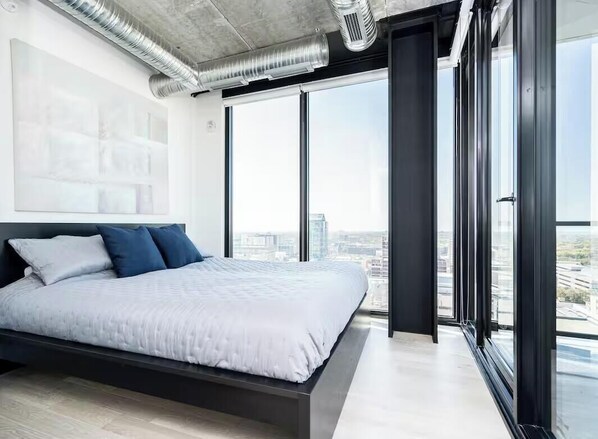 Une chambre moderne dotée d’un grand lit, offrant une vue sur la ville grâce à de grandes fenêtres du sol au plafond, et dont le système de conduits est apparent au plafond.