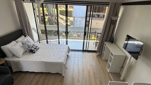 Ein Schlafzimmer mit Bett, Schreibtisch, Fernseher und einem Balkon mit Blick auf Bäume und Gebäude.