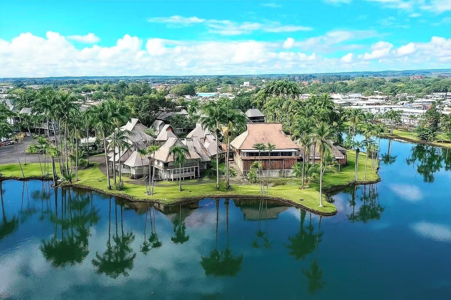 Een tropisch resort met rieten gebouwen, een centraal gebouw met een rood dak en een wateroppervlak dat de gebouwen weerspiegelt.