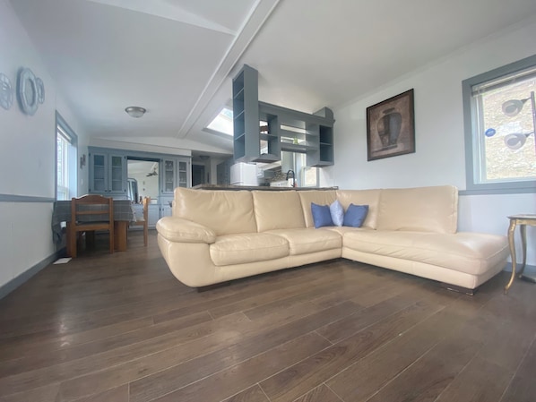 Ein Wohnzimmer mit einem beigefarbenen Ecksofa, ein Essbereich mit einem Holztisch und Stühlen sowie eine Küche mit blauen Schränken und einem weißen Kühlschrank.