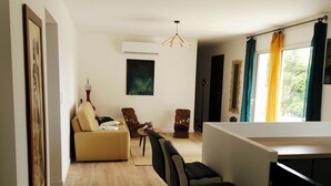 Una sala de estar moderna con un sofá, sillas, una zona de comedor con taburetes de bar, un cuadro y un ventanal con cortinas.