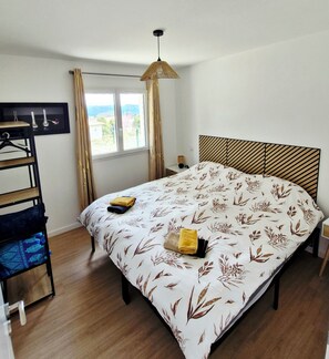 Une chambre à coucher avec un lit, une fenêtre avec des rideaux, un luminaire suspendu et une étagère fixée au mur, ornée de vases.