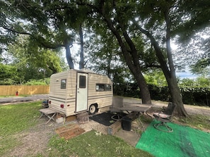 A stationary RV with a table and chairs set up in a backyard, surrounded by trees.