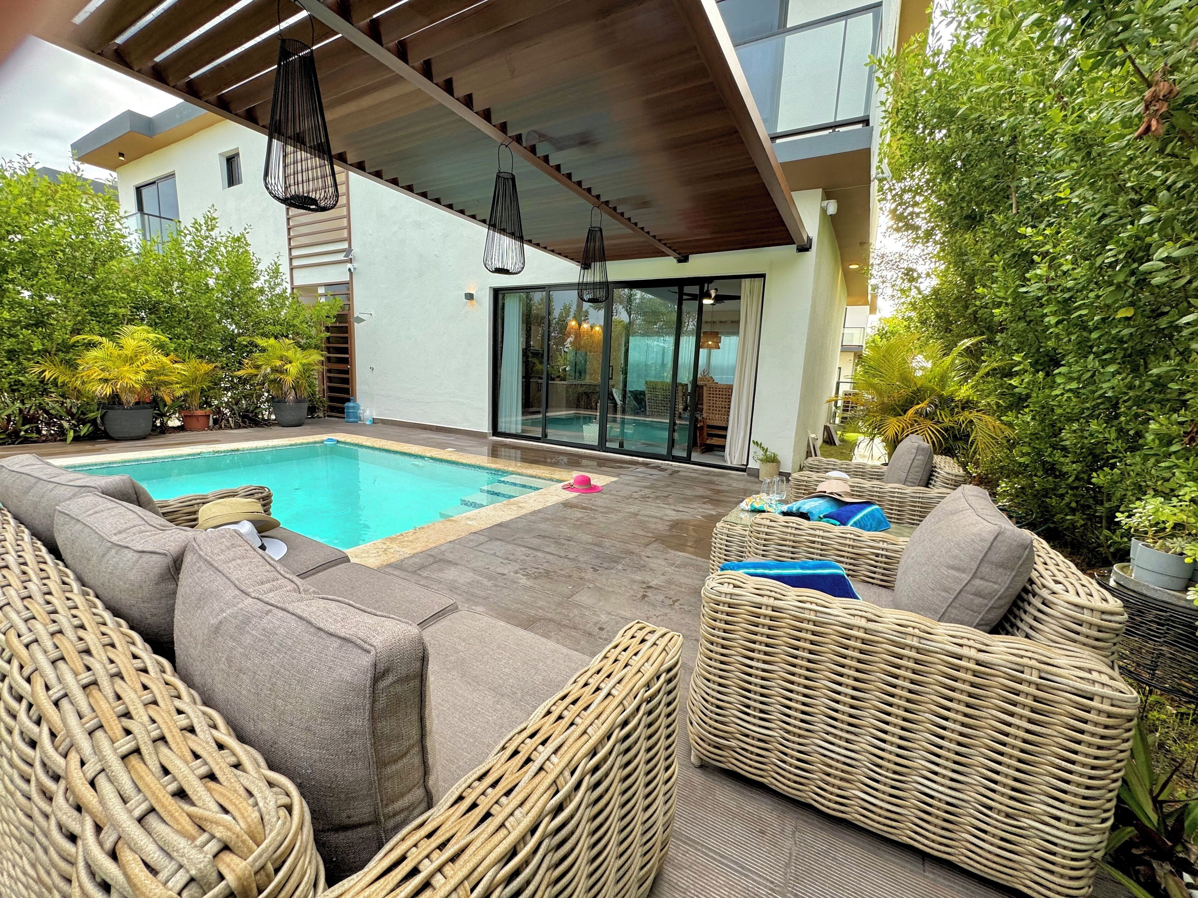 Un área de piscina moderna al aire libre con mobiliario de mimbre y una zona de estar techada.