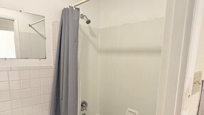 A bathroom with a glass-enclosed shower, a white tiled wall, a wooden floor, a white toilet, and a mirror.