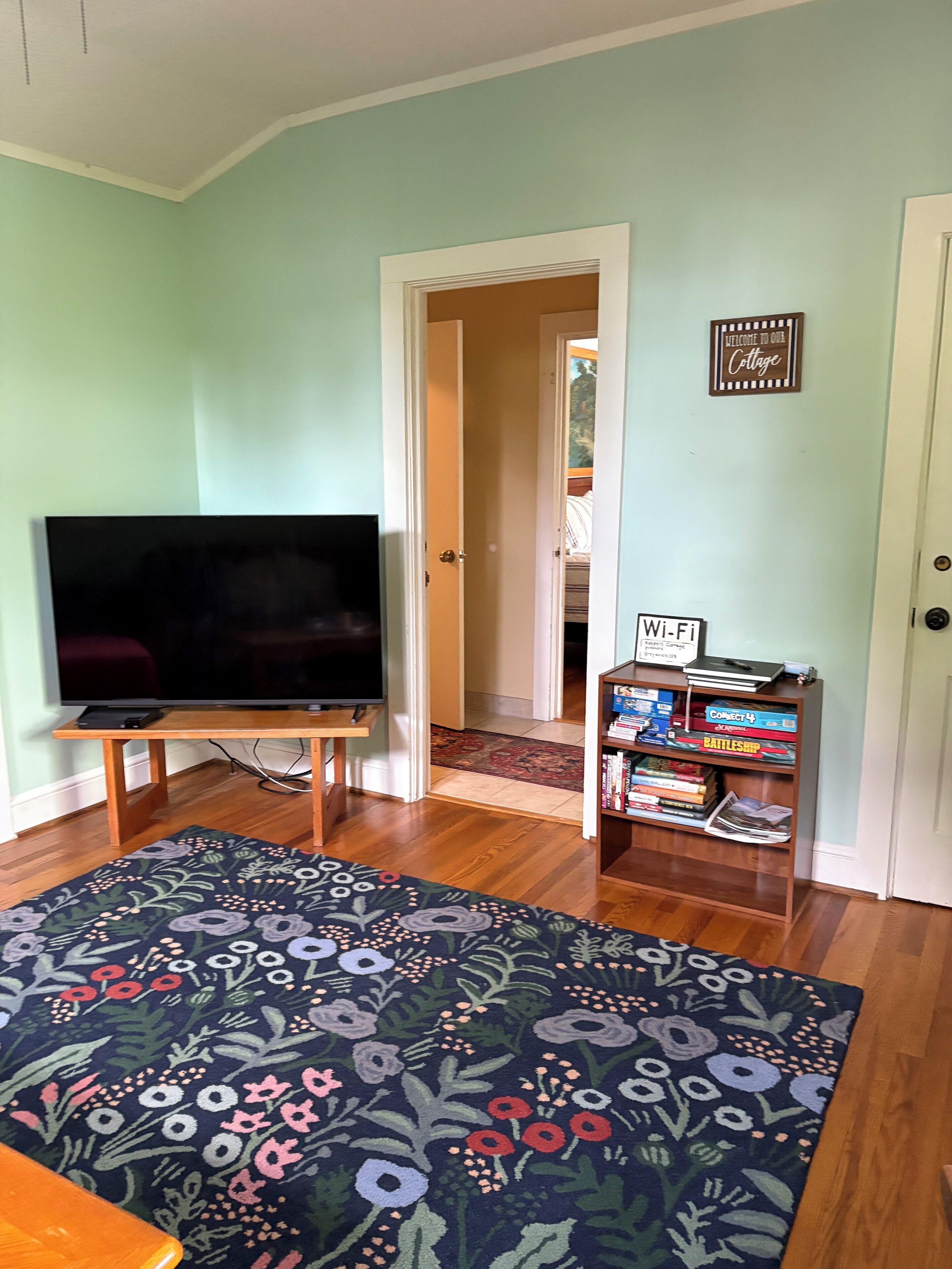 A living room with a flat-screen TV, a floral rug, a bookshelf with games, and a door leading to another room.