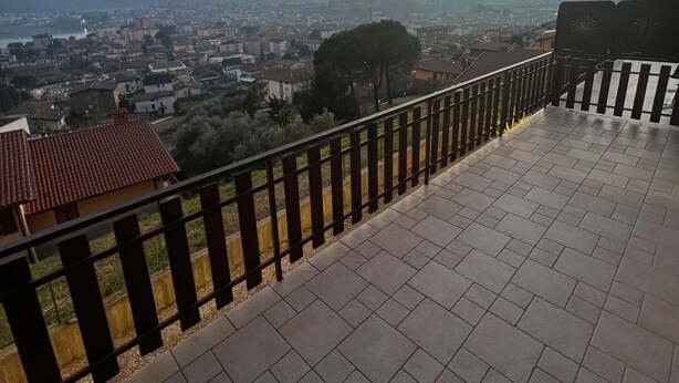 Una terrazza con pavimento piastrellato e ringhiera in metallo, affacciata su un panorama urbano con edifici e una collina all'orizzonte.