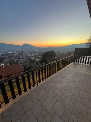 Una terraza con piso de baldosas y baranda metálica, con vista a un paisaje urbano que incluye edificios y una colina al fondo.