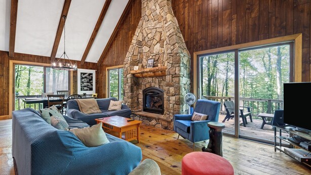 Ein gemütliches Wohnzimmer mit Kamin aus Stein, Holzbalkendecke, großen Fenstern und einer Terrassenfläche.