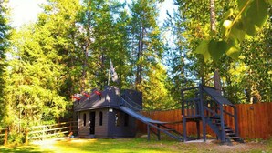 A playground with a slide and climbing structures, surrounded by trees and a wooden fence.