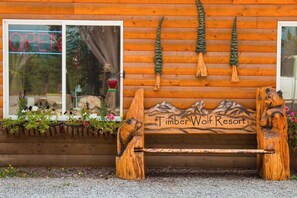 Puinen penkki, jonka selkäosassa on vuoristomainen kuvio, ja jonka edessä on puinen rakennus, jossa on ikkuna ja ikkunassa loistaa neonvaloin sana ”AVOIN”. Penkin edessä on myös teksti ”Timber Wolf Resort”.