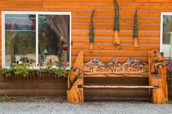 Un banco de madera con la inscripción 'Timber Wolf Resort', una ventana con un cartel de 'ABIERTO' y una pared de madera con adornos colgantes.