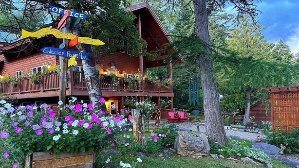 Una cabaña de madera con un porche, rodeada por un jardín con flores rosas y blancas, y un cartel con indicaciones.