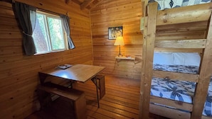 Interior de cabaña con revestimiento de madera, una litera, un escritorio, una lámpara y una ventana con cortinas.