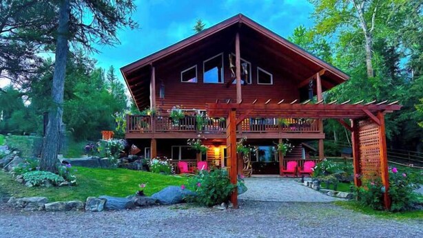 A two-story wooden cabin with a balcony, surrounded by a well-maintained garden and trees. - Cute Camper on FHU Site in Beautiful Campground (Hungry Horse)