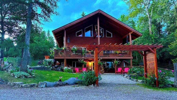 A two-story wooden cabin with a balcony, surrounded by a well-maintained garden and trees. - Lodge Room with Views of Glacier NP (Hungry Horse)