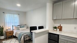 Un apartamento moderno y compacto con kitchenette, una cama con cabecera, una pequeña zona de comedor y una planta en maceta.