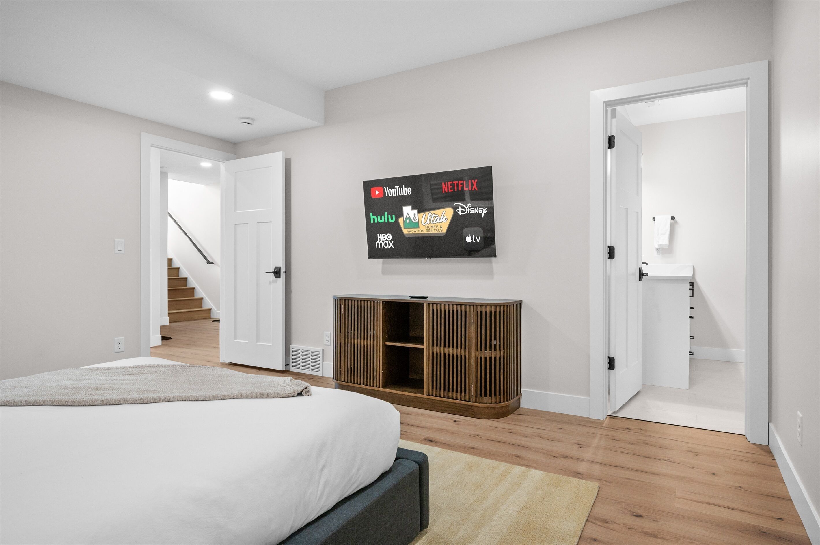 Un dormitorio moderno con una cama grande, un televisor montado en la pared mostrando varias plataformas de streaming y un mueble de madera debajo.