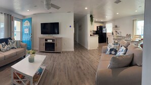 A modern living room with a beige sofa, a white coffee table, and a TV. There's a kitchen area with a black refrigerator and a dining table with chairs.