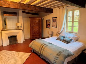 A bedroom with a large bed, a wooden wardrobe, a fireplace, and a window with a view of greenery.