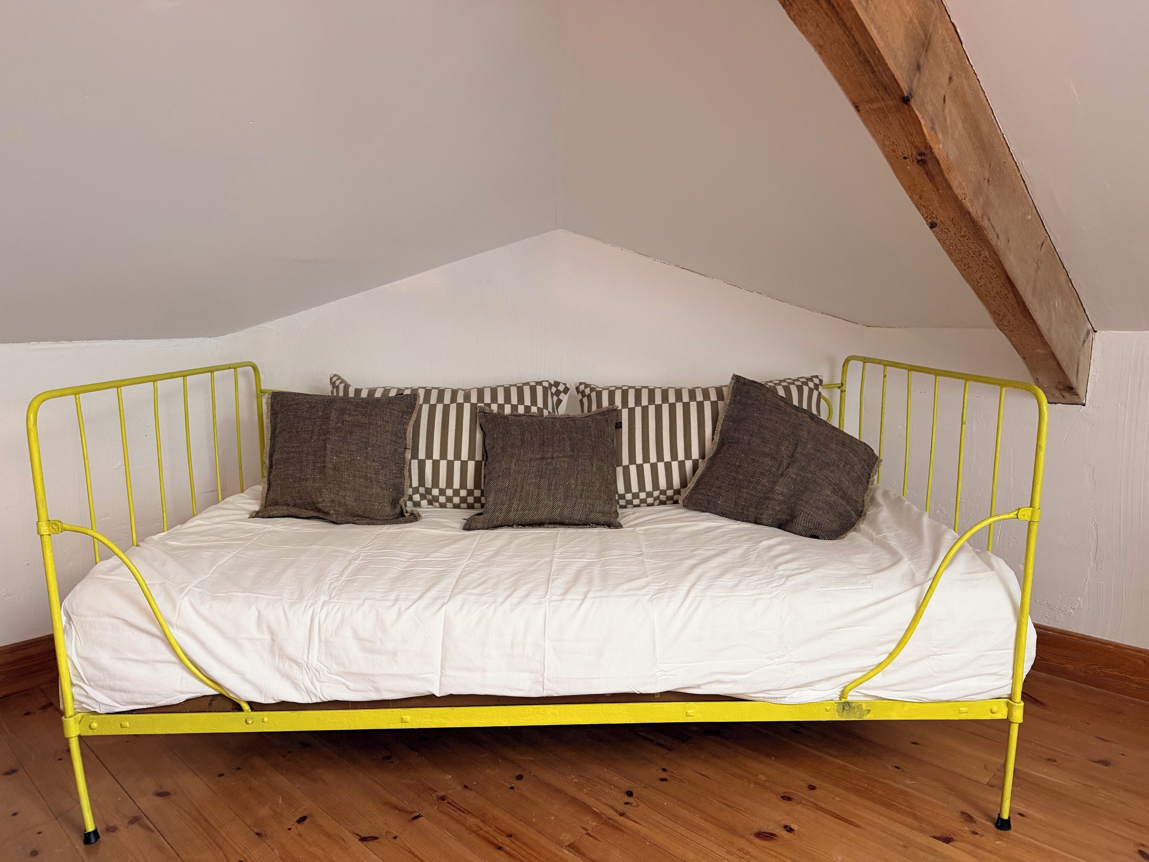 Un lit en métal jaune, avec une literie blanche et plusieurs oreillers, dans une pièce au plancher en bois et au plafond en pente.