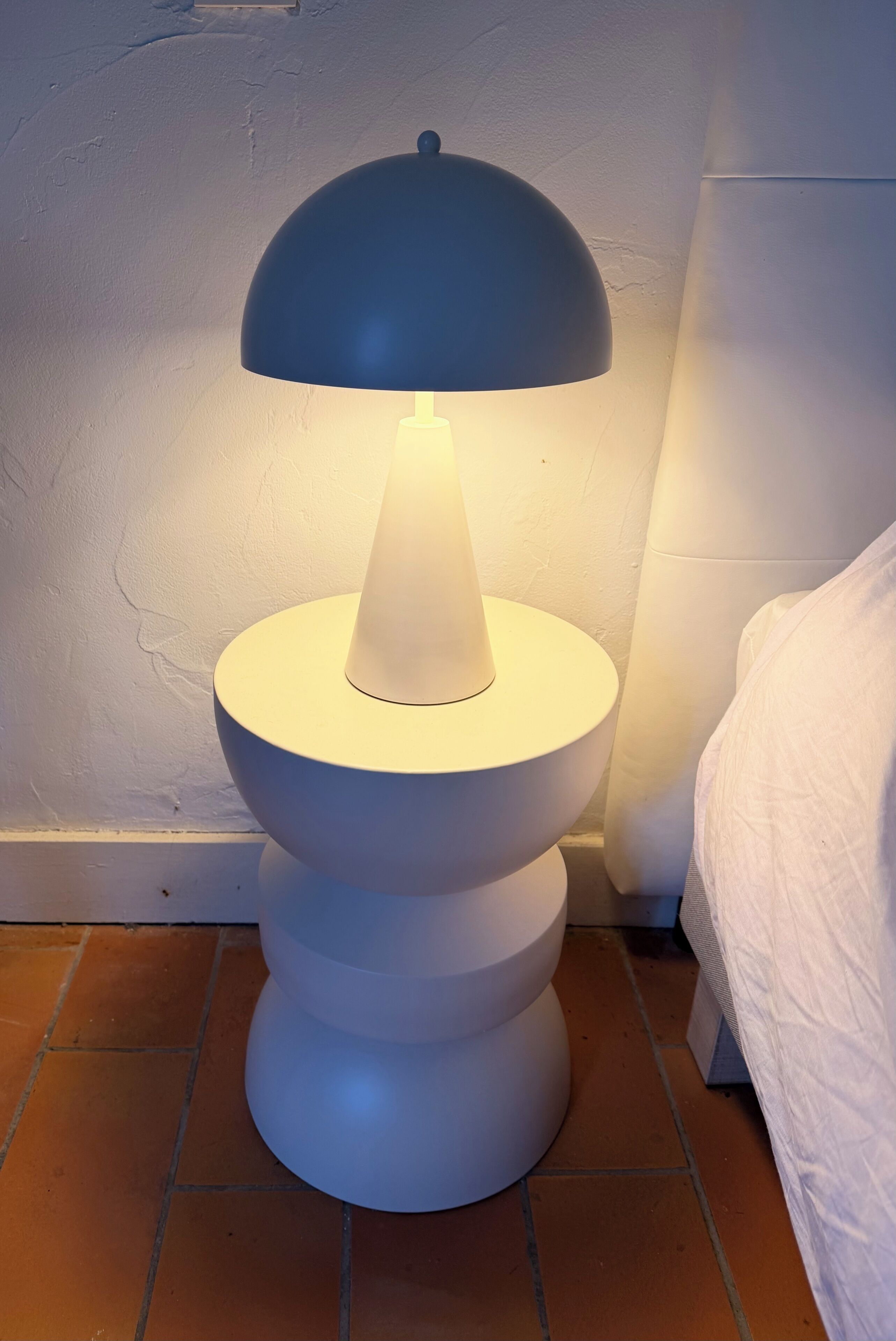 A modern floor lamp with a white base and a blue shade, positioned on a tiled floor next to a bed with white bedding.