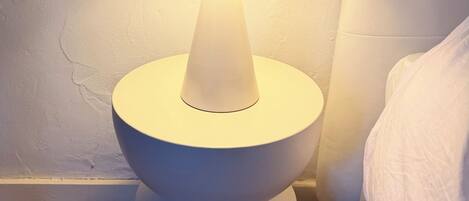 A modern floor lamp with a white base and a blue shade, positioned on a tiled floor next to a bed with white bedding.