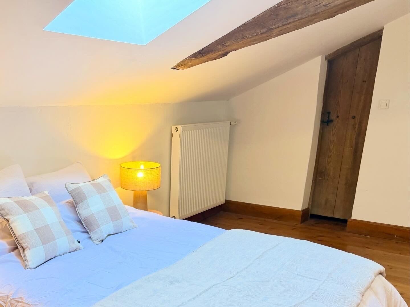 Ein Schlafzimmer mit Dachfenster, einem Bett mit Karokissen, einer Nachttischlampe, einem Heizkörper und einer Holztür.