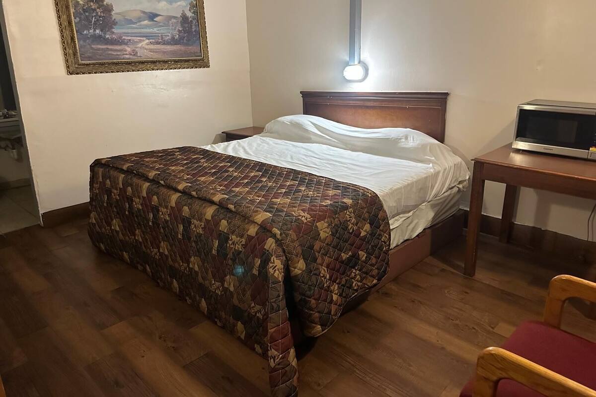 Ein Schlafzimmer mit einem Bett, einem Nachttisch aus Holz, einem Beistelltisch mit einer Mikrowelle, einem Sessel, einem Bild an der Wand und Holzboden.