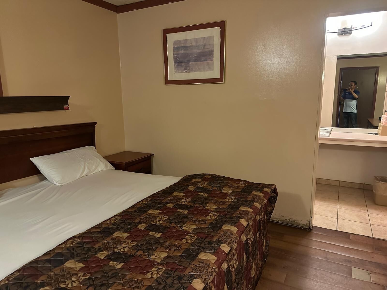 A hotel room with a bed, a nightstand, a framed picture on the wall, and a bathroom visible through an open door.