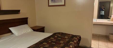A hotel room with a bed, a nightstand, a framed picture on the wall, and a bathroom visible through an open door.