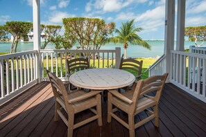 Eine Holzterrasse mit einem runden Tisch und vier Stühlen, mit Blick auf das klare, blaue Meer und eine Rasenfläche mit Bäumen und einer Palme.