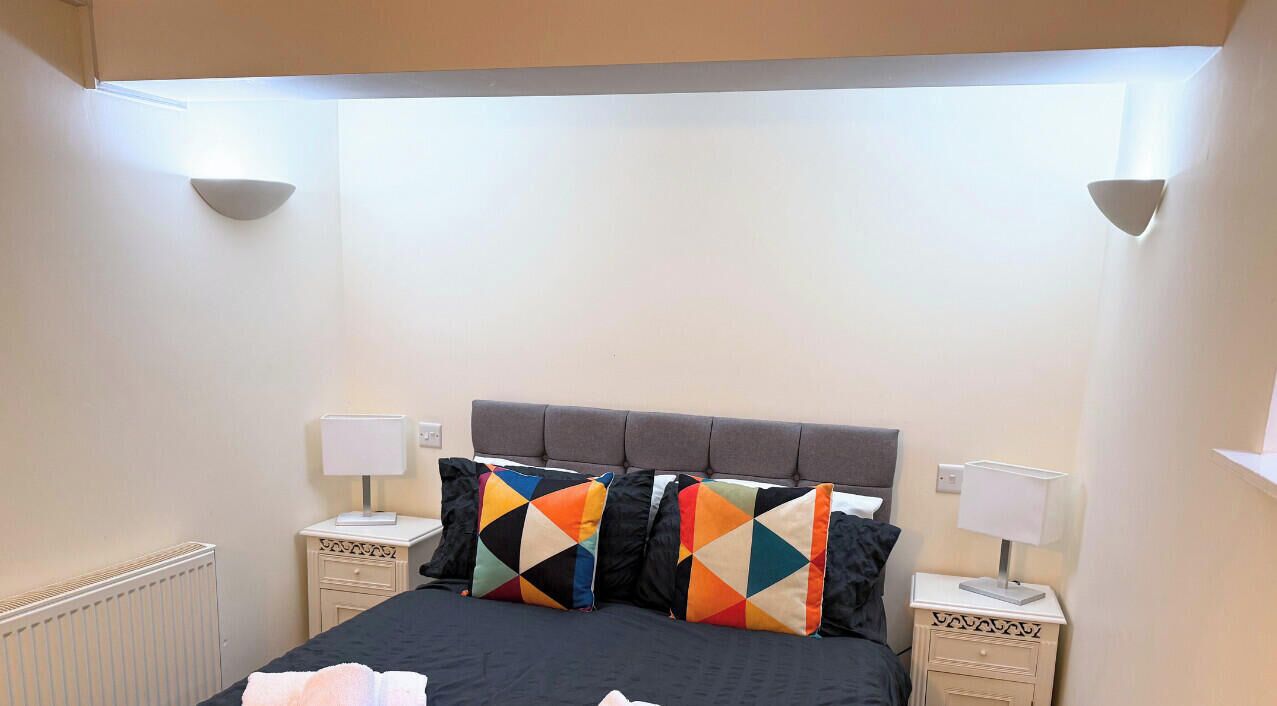 Een net opgemaakt bed met een zwart dekbed, twee kussens met kleurrijke geometrische patronen, en twee witte nachtkastjes met lampen.