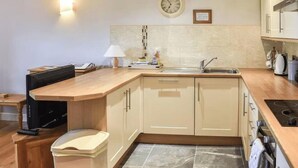 A modern kitchen with a wooden countertop, a sink, and a stove. There is a clock on the wall and a small table with a lamp. - Crag Barn Cottage – Lake District Getaway by PolkaStays (Lake District)