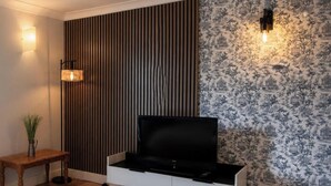 A modern living room with a flat-screen TV, a wooden slat wall, a patterned wallpaper, a standing lamp, and a small table with a plant. - Crag Barn Cottage – Lake District Getaway by PolkaStays (Lake District)