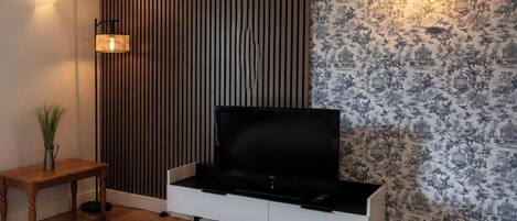 Ein modernes Wohnzimmer mit Flachbildfernseher, einer Wand aus Holzleisten, gemustertem Tapeten, einer Stehlampe und einem kleinen Tisch mit einer Pflanze.