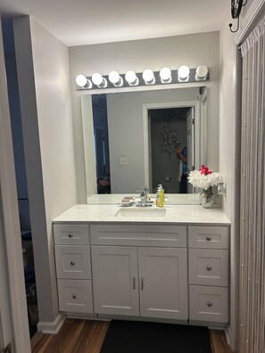 A bathroom with a vanity, a mirror with lights, a sink, and a vase with flowers.