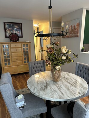 A dining area with a marble table, grey upholstered chairs, a vase with flowers, a chandelier, and a wooden cabinet with glass doors.