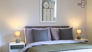 A neatly made bed with a white and green bedspread, two grey pillows, and a headboard. There are two bedside lamps on white nightstands. A framed artwork with the text "OH HI" is on the wall above the bed. - The Stables | Romantic Countryside Escape in the Lake District (Lake District)