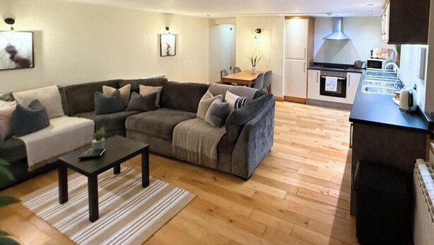 A modern living room with a sofa, coffee table, and a dining table with chairs. There is a kitchen area with a stove and microwave. - The Stables | Romantic Countryside Escape in the Lake District (Lake District)
