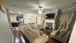 Een woonkamer met een grote beige hoekbank, een kleinere fauteuil en een open haard. Achtergrond is een keuken met een eettafel en stoelen.