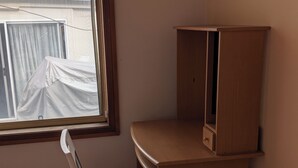 Ein Eckzimmer mit Schreibtisch und Stuhl aus Holz, einem Fenster mit Vorhang und Blick auf ein Gebäude.