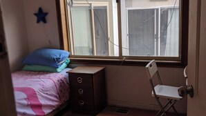 Ein Schlafzimmer mit einem Einzelbett, einem kleinen Schminktisch, einem Stuhl und einem Fenster mit Blick auf Gebäude.