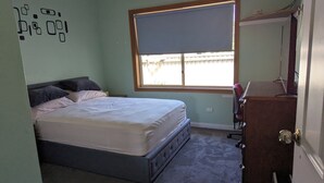 Ein Schlafzimmer mit Bett, Kommode, Schreibtisch und einem Fenster mit Jalousie.