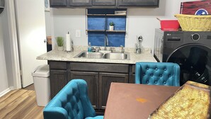 A compact kitchen with dark wooden cabinets, a granite countertop, a sink, a washing machine, and a blue upholstered chair. - Deck the Halls (Clarksville)