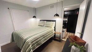A bedroom with a bed, bedside tables, a nightstand, a ceiling light, a wall-mounted heater, and a door leading to a kitchen. - Deck the Halls (Clarksville)