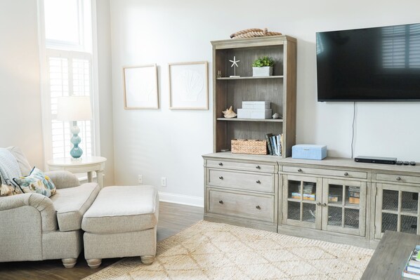 A living room with a sofa, ottoman, wooden cabinet, flat-screen TV, and a small table with a lamp. - Large beach home w golf cart, minutes from beach! (St Simons Island)