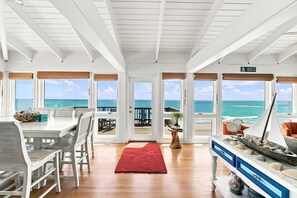 Ein geräumiges Zimmer mit Holzboden, einer Essecke mit Tisch und Stühlen, einem roten Teppich und großen Fenstern mit Blick auf den Ozean.