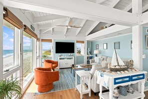A coastal living room with a large window, a flat-screen TV, and a dining area with a table set for two. - Bonsai - Oceanfront Zen Retreat • 4BR • 5BA • Direct Beach Walkway • Epic Views (Oak Island)